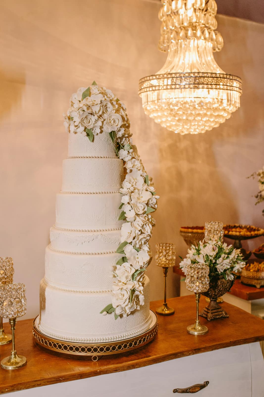 A tiered wedding cake dressed with flowers beneath a chandelier